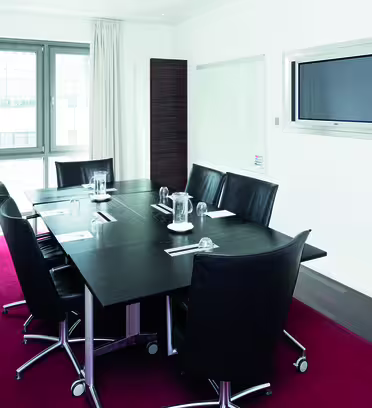 A modern conference room with a rectangular black table surrounded by six black leather chairs. The room has white walls, white curtains, a red carpet, and a flat-screen TV mounted on the wall.