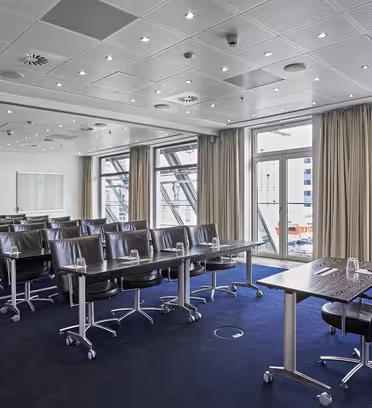 Conference room with rows of black leather swivel chairs and tables, each set with glasses and paper, facing a large window overlooking a cityscape. Ceiling lights and neutral decor create a professional atmosphere.