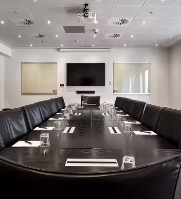 A modern conference room with a long, dark wooden table surrounded by black leather chairs. Notepads and glasses are placed at each seat. The room has a large screen on the wall, bright lighting, and a window with curtains on the right side.