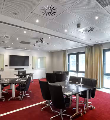 Meeting room with red carpet, two glass tables, and black chairs. A lectern and large screen are positioned at the front. Large windows and bright overhead lighting illuminate the space.