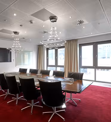 A modern conference room with a long wooden table surrounded by black chairs on a red carpet. Two large windows provide natural light, and two ceiling light fixtures illuminate the space. Beige curtains hang next to the windows.