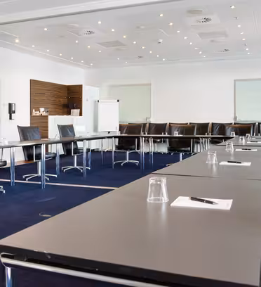 A modern meeting room with a U-shaped arrangement of black chairs and tables. Each table has a notepad, pen, and glass. The room has bright lighting and a flip chart at the front. White walls and a dark carpet complete the setting.