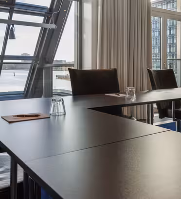 A modern conference room with large windows showcases a waterfront view. Black tables form a U-shape, each with a notepad, pen, and an upside-down water glass. Beige curtains frame the windows, and theres a blue carpet on the floor.