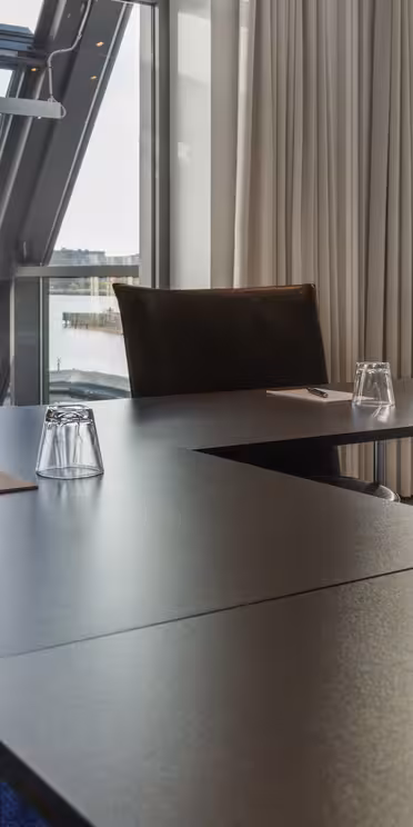 A modern conference room with large windows showcases a waterfront view. Black tables form a U-shape, each with a notepad, pen, and an upside-down water glass. Beige curtains frame the windows, and theres a blue carpet on the floor.