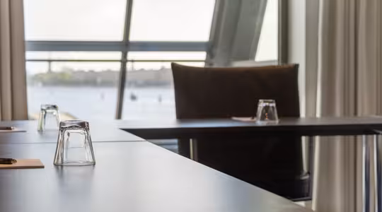 Modern conference room with glass walls, offering a view of a waterfront. Tables are set with upside-down glasses and notepads. A large chair is seen, with light streaming through curtains.