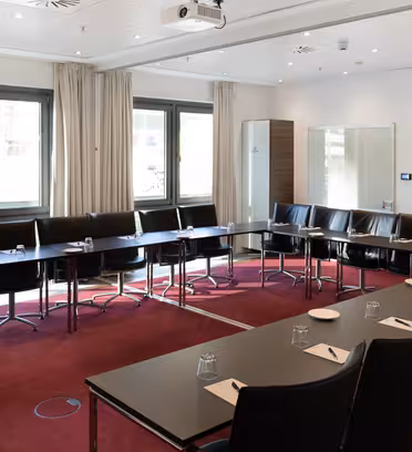 A modern conference room with tables arranged in a U-shape, surrounded by black chairs. Each table has a notepad, pen, and glass of water. Theres a flat-screen TV on the wall, a projector on the ceiling, and large windows with beige curtains.