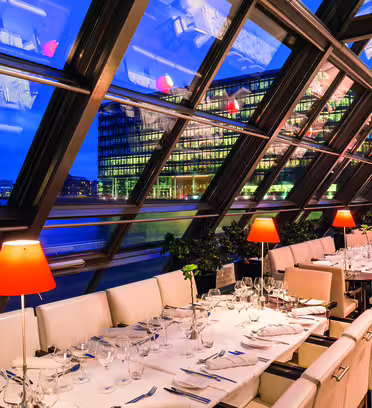 A modern restaurant with floor-to-ceiling angled windows offers a view of a cityscape at dusk. Tables are set with white cloths, glassware, and cutlery. Red lamps add a warm glow. City lights are visible outside.