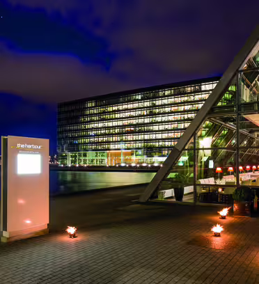 Modern waterfront building with glass facade at night, featuring lit interiors and reflections on the water. An illuminated sign reads the harbour, and ground lights enhance the ambiance.