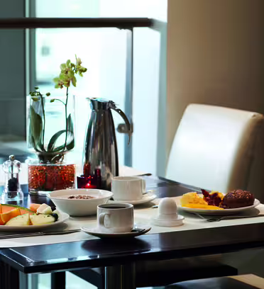 A table set for breakfast with a plate of fruit, cheese, eggs, and bread. A cup of coffee, a small pitcher, and a plant centerpiece are also on the table. The setting is in a bright, modern dining space.