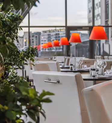 A modern restaurant with white chairs and tables set with glassware. Red lamps line the tables on the right side. Large windows reveal an urban cityscape outside, and leafy plants are visible in the foreground.