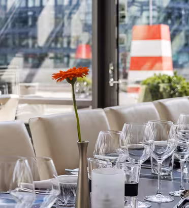 A restaurant table is set with empty wine glasses, water glasses, and white napkins. A single orange flower in a vase decorates the center, with large windows and a blurred cityscape in the background.
