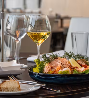 A table set with a dish of smoked salmon garnished with dill and lemon slices, served on a bed of lettuce. Two glasses of white wine, a pepper grinder, and a plate of sliced bread with butter accompany the dish.