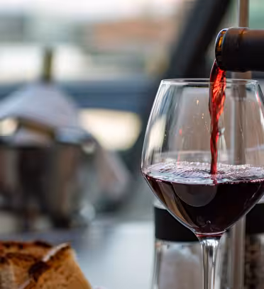 A glass of red wine is being poured from a bottle on a dining table. In the background, theres a blurred view of a bottle in an ice bucket. Some bread slices and salt and pepper shakers are visible.