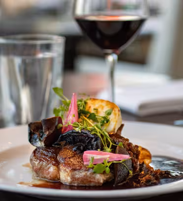 A gourmet dish of steak with vegetables, garnished with herbs and purple onion slices, served on a white plate. Theres a glass of red wine in the background, along with a water glass and a salt shaker on the table.