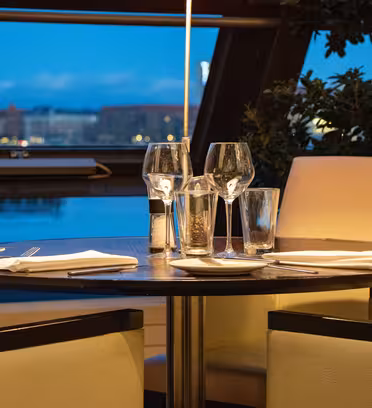 Elegant restaurant table set for four with wine glasses, napkins, and cutlery. Two beige cushioned chairs are visible. Large windows reveal a cityscape with buildings under a blue evening sky, adding a sophisticated ambiance.