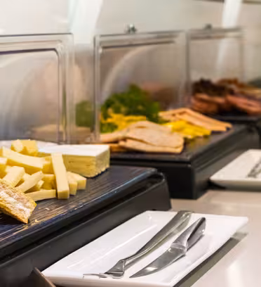 An assortment of cheeses and meats are displayed on a buffet table. Slices of various cheeses are in the foreground, with meats and greens under clear lids in the background. Plates and cutlery are set up nearby.