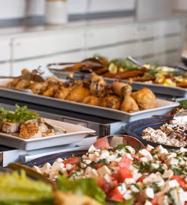 A buffet table filled with various dishes, including salads, roasted chicken drumsticks, pasta, and garnished meats. Fresh greens and herbs are used as garnishes. Plates and utensils are visible in the background.
