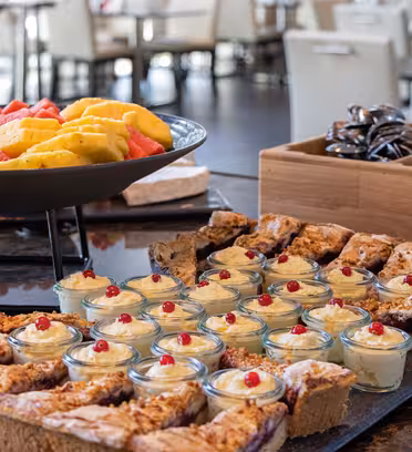 A variety of desserts are displayed on a table, including fruit slices in a black bowl, small glass dishes of cream topped with red berries, and assorted pastries on a tray. A wooden box holds wrapped chocolates. Background shows a dining area.
