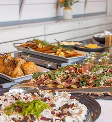 A buffet table filled with dishes including roasted chicken, cold cuts with greens and pickles, a mixed salad, and assorted breads. The setting appears elegant, with modern decor and a floral arrangement in the background.
