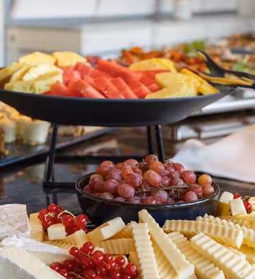 A table filled with a variety of foods, featuring a tray of sliced watermelon and pineapple, a bowl of red grapes, assorted cheeses, and desserts like fruit tarts. The setting suggests a buffet-style spread.