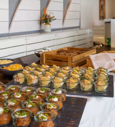 A buffet table with a variety of foods, including small glass jars filled with salads and meats, a wooden tray with bread rolls, and a bowl of lemon wedges. The table is set in a modern dining area with decorative elements.