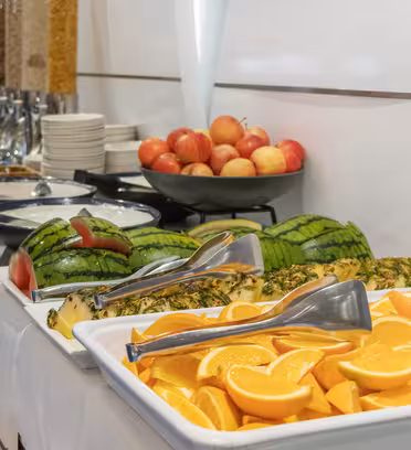 A buffet table with sliced oranges and pineapples, watermelon pieces, a bowl of apples, pitchers of cereal, and stacks of plates. Tongs are available for serving.