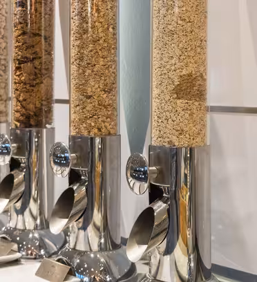 Five transparent cereal dispensers in a row, each filled with different types of cereal. The dispensers have metallic bases with levers for dispensing.