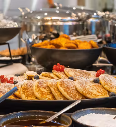 A plate of pancakes topped with blueberries and red currants, dusted with powdered sugar, is in the foreground. In the background, there are bowls of food and various utensils on a buffet table.