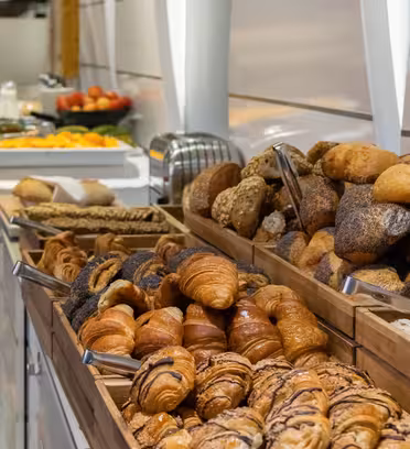 A buffet table lined with various breads and pastries in wooden trays, including croissants and rolls. In the background, theres a platter with sliced fruits and some stacked plates. The setting appears to be a breakfast buffet.