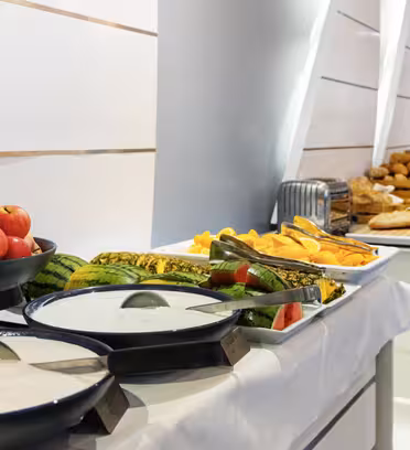 A buffet table with bowls of yogurt, plates of sliced fruit, apples, and an assortment of bread and pastries. The table is set in a modern room with sleek, white decor.