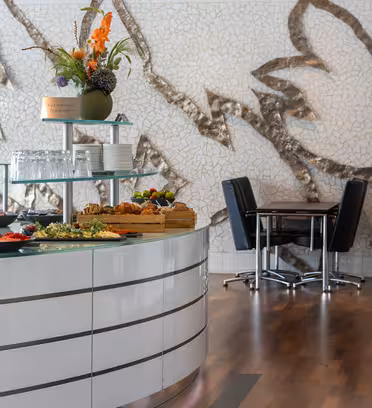 A modern dining area with a buffet table featuring assorted pastries, fruit, and salads. The backdrop is a large mosaic art piece, and there are black leather chairs around tables with a wooden floor.