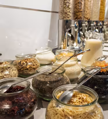 A breakfast buffet table with jars of various cereals, nuts, dried fruits, and seeds. Dispensers with cereal and a jug of milk are visible in the background. Spoons are placed in the jars for self-service.
