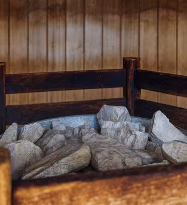 A metal container filled with large, uneven rocks is placed within a dark wooden frame. The background consists of vertical wooden panels, giving the setting a rustic sauna-like appearance.