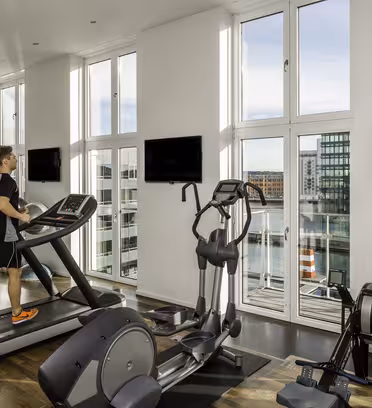 A person is running on a treadmill in a modern gym with large windows showing a cityscape and water view. The gym features exercise equipment and two wall-mounted TVs. Bright natural light fills the space.