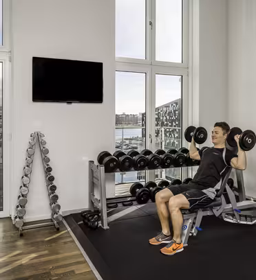 A person exercises in a bright gym with large windows overlooking a cityscape. They are seated on a bench, lifting dumbbells. Nearby, there are racks with more weights, and a wall-mounted TV. The floor is a mix of wood and rubber matting.