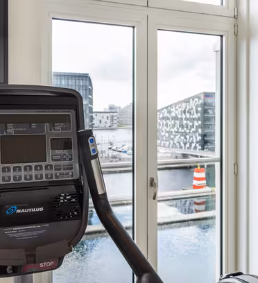 View from a modern exercise bike placed indoors, facing a large window. The window overlooks a waterfront with industrial-style buildings and a buoy visible outside. A flat-screen TV is mounted on the wall to the right.