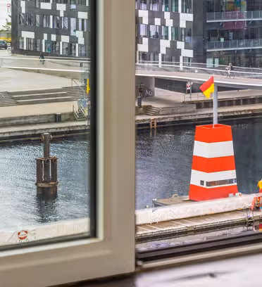 View from a window overlooking a canal with a large red and white buoy. Buildings and a walkway with people and parked cars are visible in the background. The scene is urban and modern.
