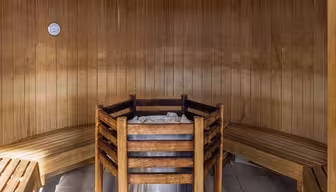 Interior of a wooden sauna featuring a central heater surrounded by a wooden guardrail. Two curved wooden benches line the walls, and a small thermometer is mounted on the wooden paneling. The floor is tiled.