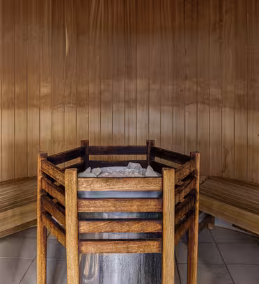 Interior of a wooden sauna featuring a central heater surrounded by a wooden guardrail. Two curved wooden benches line the walls, and a small thermometer is mounted on the wooden paneling. The floor is tiled.