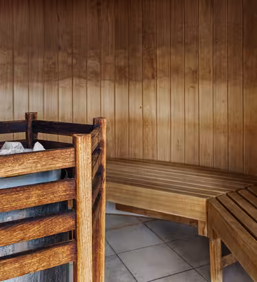 A wooden sauna interior with curved benches lining the wall. A bucket with a ladle sits on a bench, and a heater filled with stones is on the left. The warm wood tones create a cozy atmosphere.